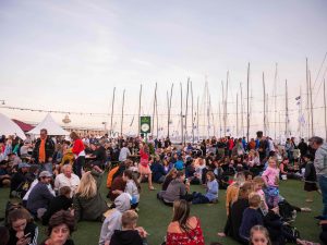 image of people sitting on fake grass area in Sydney Hobart Yacht Race Village as the sun sets behind all the yachts in background