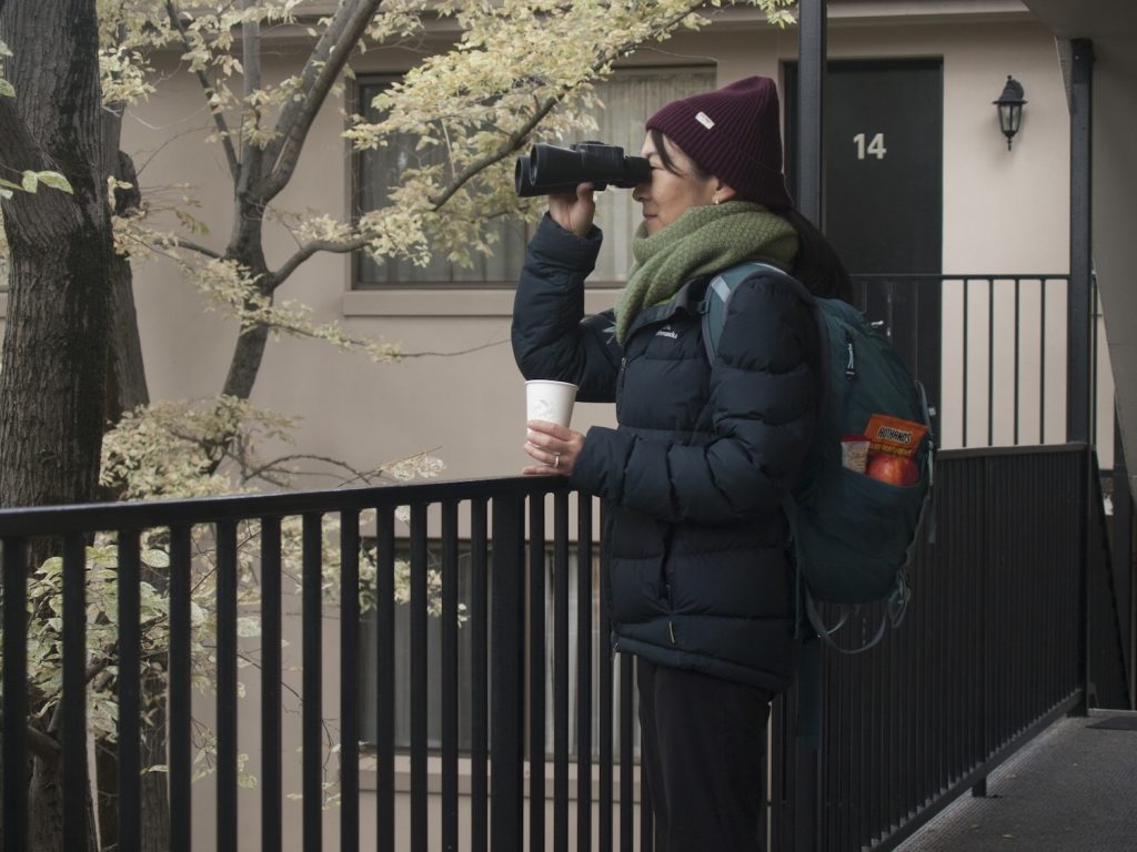 Guest rugged up in winter clothes. Holding a coffee and pair of binoculars.